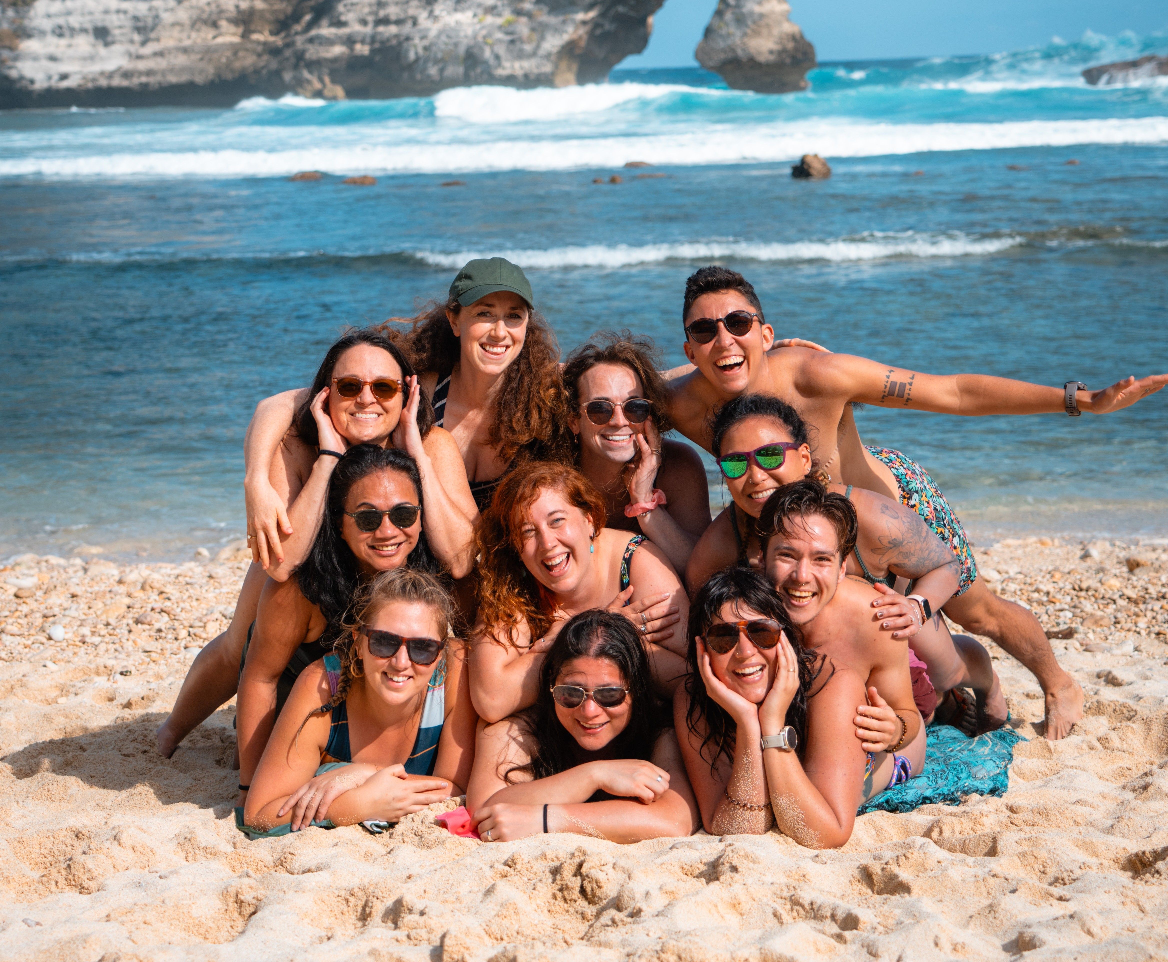 A group of smiling friends poses closely together on a sandy beach with the ocean behind them.