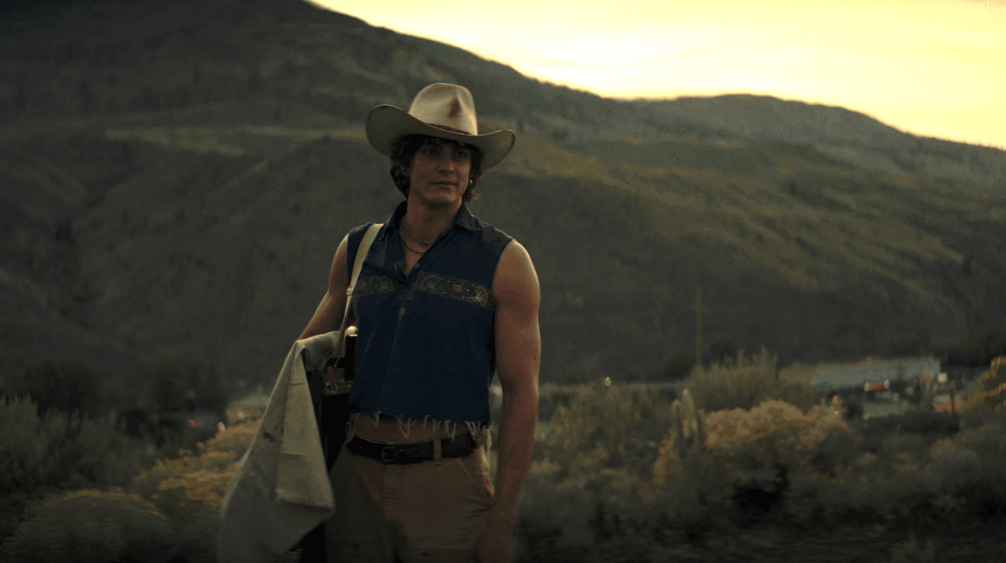 A man in a cowboy hat and denim vest stands confidently in a desert at sunset.